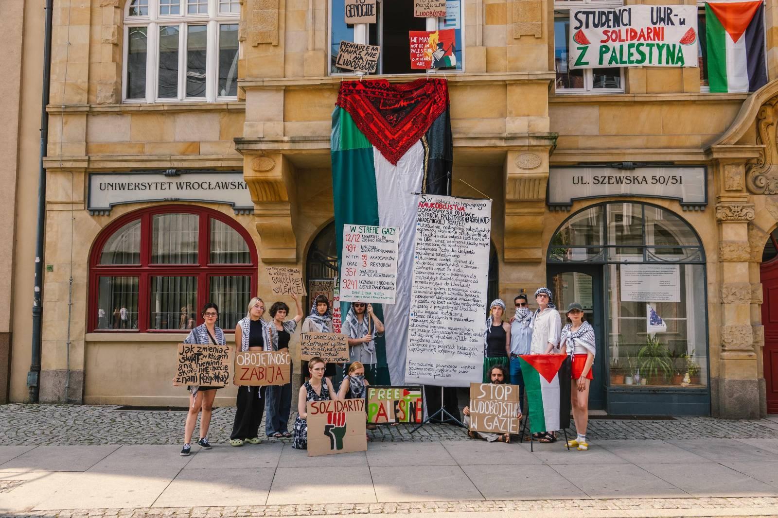 Konferencja prasowa, na której studenci ogłosili piąty postulat. Stoją przed budynkiem Instytutu Kulturoznawstwa z banerami i treścią postulatu. Z okna naad nimi wisi flaga Palestyny.