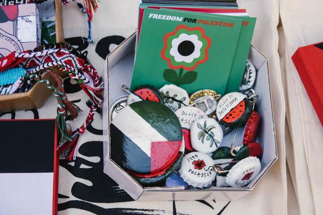 Small box with pro-Palestinian pins and stickers sold at the fair. Next to them, in the shot, there are hand-made bracelets.