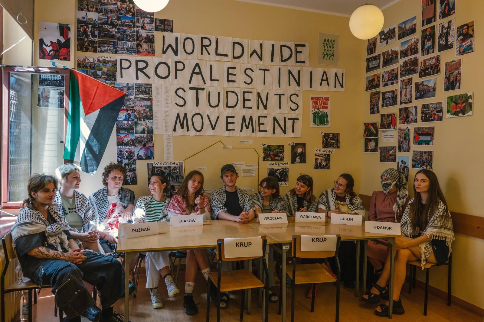 W okupowanej sali siedzą studenci z polskich okupacji. Nad nimi na ścianach przyklejone są zdjęcia z protestów i napis “Worldwide Propalestinian Students’ Movement”.