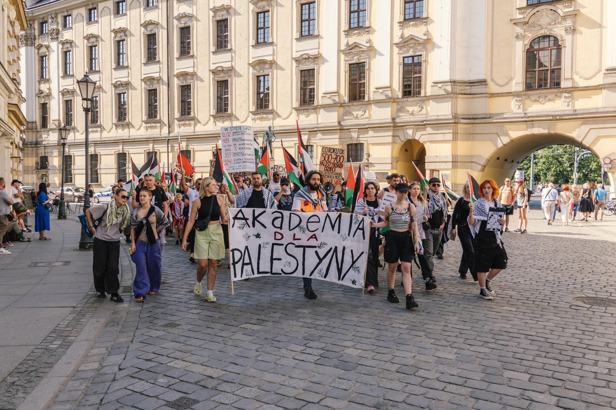 Osoby demonstrujące rozpoczynające marsz na Placu Uniwersyteckim pod fontanną z Szermierzem. Osoby z przodu niosą baner „Akademia dla Palestyny”, za nimi widać gmach główny Uniwersytetu.